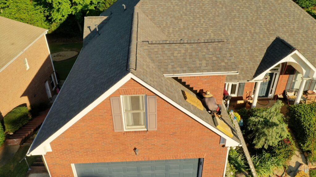 A construction worker in a straw hat is repairing shingles on the roof of a brick house, using a ladder for access.